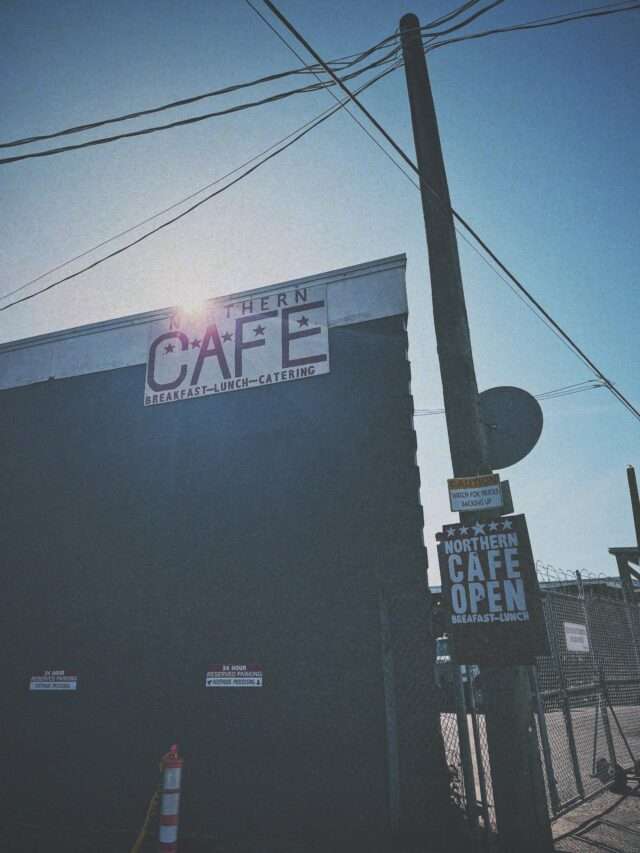 Street corner with a large cafe sign reading "Northern Cafe Open" against a clear blue sky and utility pole with wires overhead (urban storefront scene).