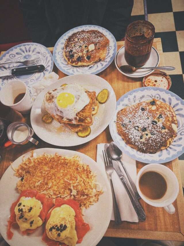 Overhead view of a brunch table with plates of pancakes, fried eggs, and pastries, plus coffee cups and a jam jar.