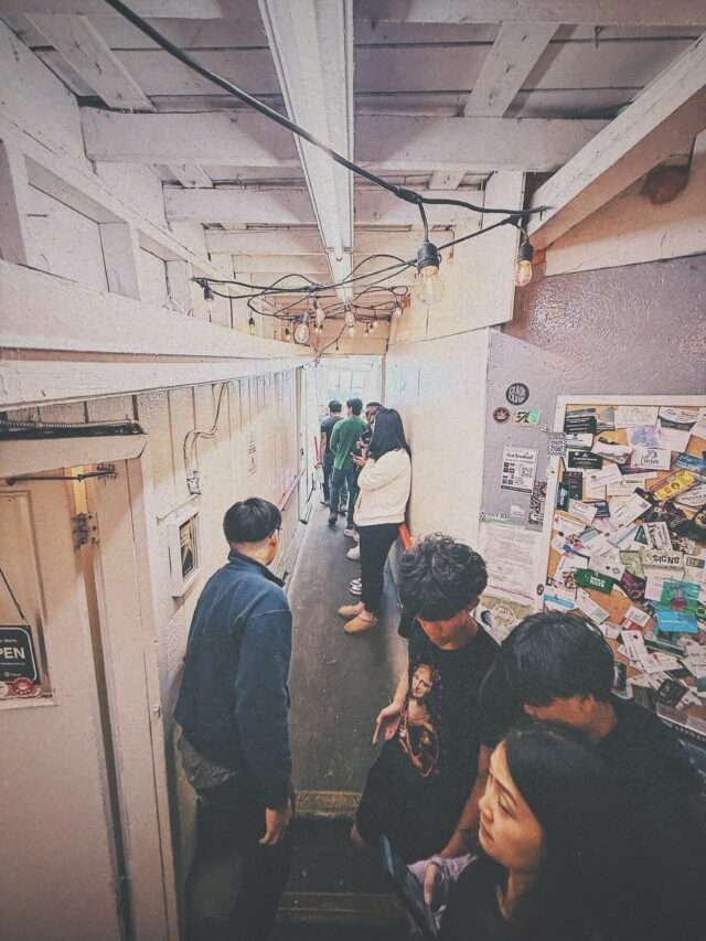 Line of people standing in a narrow, dim corridor with bare wooden beams, exposed wiring, and hanging light bulbs; a bulletin board covered with stickers on the wall.